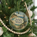 A round ornament hanging on a Christmas tree, featuring a scenic landscape with a lake and mountains.