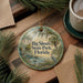 A circular ornament with a scenic landscape of Big Shoals State Park, Florida, featuring a bird and palm trees, is displayed on a wooden surface alongside a cup of coffee and some pine branches.