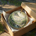A round ornament in a box, featuring a scenic landscape with a waterfall and trees, with the text "Bigelow Hollow State Park Connecticut" printed on it.