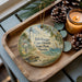 A decorative ornament featuring a lighthouse and beach scene, with the text "Bill Baggs Cape Florida State Park, Florida" printed on it. The ornament is placed on a wooden tray, surrounded by pine cones and a lit candle.