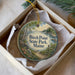 A round ornament in a box, featuring a scenic landscape with a path leading to a body of water and trees.