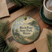 A circular ornament with a scenic landscape of Birch Point State Park, Maine, featuring a path, trees, and water, is displayed on a wooden surface alongside a cup of coffee and some pine branches.