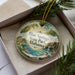 A decorative ornament with a scenic image of Blue Springs State Park, Alabama, displayed in a box with pine branches.