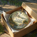 A decorative ornament in a brown box, featuring a scenic landscape with a castle, trees, and a river.