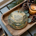 A wooden tray with a decorative ornament featuring a scenic mountain landscape, pine cones, and a lit candle.