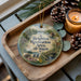 A wooden tray with a decorative ornament featuring a scenic landscape and a deer, along with pine cones and a lit candle.