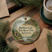A decorative ornament featuring a scenic lake and resort park in Kentucky, with a boat on the water and a cabin in the background. The ornament is hanging from a string and is placed on a wooden surface, surrounded by pine branches and a cup of coffee.