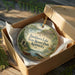 A decorative ornament in a box, featuring a scenic landscape with a lake, mountains, and a cabin, along with the text "Buckhorn Lake State Resort Park, Kentucky".