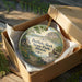 A decorative ornament in a wooden box, featuring a scenic landscape with a house, trees, and a body of water.