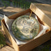 A circular ornament with a painting of a historical site, placed in a cardboard box with a gold string.