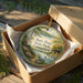 A decorative ornament in a box, featuring a scenic landscape with a lake, trees, and a cabin, with the text "Burr Pond State Park, Connecticut" printed on it.