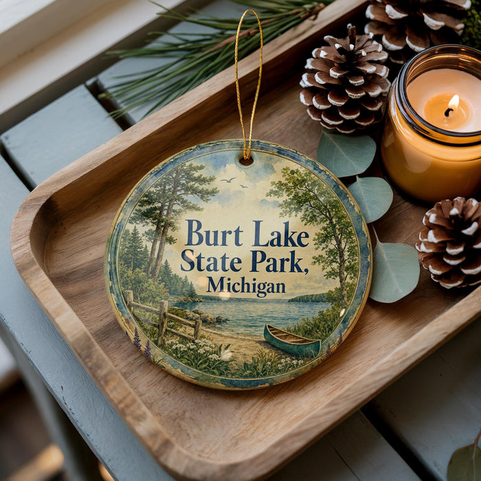 A decorative ornament with a scenic image of Burt Lake State Park, Michigan, is displayed on a wooden tray alongside pine cones and a lit candle.