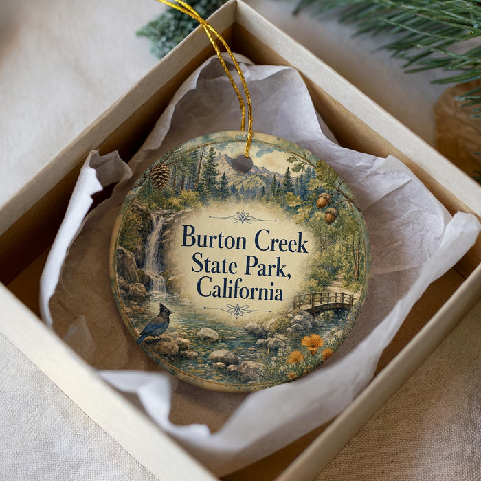 A decorative ornament in a white box, featuring a scenic landscape with a waterfall, pine trees, and a bird.