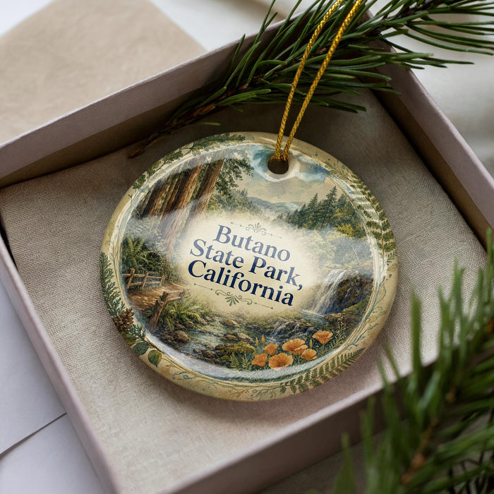 A decorative ornament with a landscape scene featuring a waterfall and pine trees, placed inside a box with a pine branch.