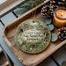 A decorative ornament with a scenic image of the Carter Caves State Resort Park in Kentucky, featuring a bridge and pine cones, placed on a wooden tray alongside a lit candle.