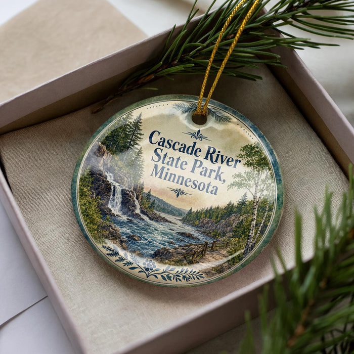 A decorative ornament in a box, featuring a scenic landscape with a waterfall and pine trees.