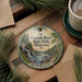 A decorative ornament featuring a scenic image of the Cascade River in Minnesota, with pine trees and a waterfall, is displayed on a wooden surface alongside a cup of coffee and some papers.