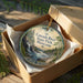 A round ornament in a box, featuring a scenic image of the Cascade River in Minnesota.