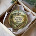 A decorative ornament in a box, featuring a scenic landscape with a castle, trees, and a river.