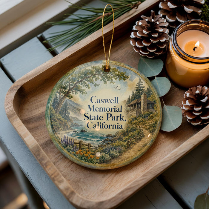 A decorative ornament with a scenic landscape of Caswell Memorial State Park, California, featuring a winding path, pine trees, and a small cabin. The ornament is displayed on a wooden tray, with pine cones and a lit candle nearby.