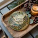 A decorative ornament with a scenic landscape of Caswell Memorial State Park, California, featuring a winding path, pine trees, and a small cabin. The ornament is displayed on a wooden tray, with pine cones and a lit candle nearby.