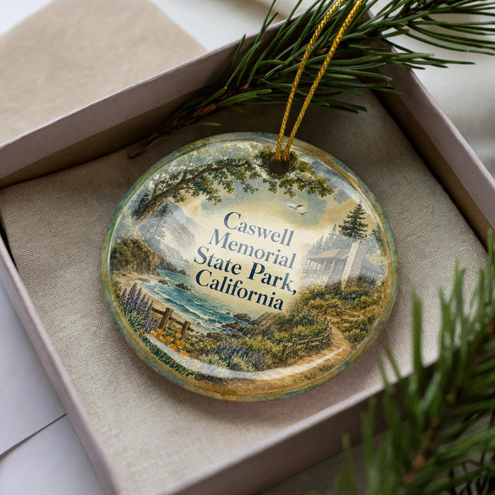 A decorative ornament in a box, featuring a scenic landscape with a house, trees, and a body of water.