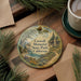 A decorative ornament with a scenic landscape of Caswell Memorial State Park, California, featuring a cabin and a river, is displayed on a wooden surface alongside a cup of coffee and some pine branches.