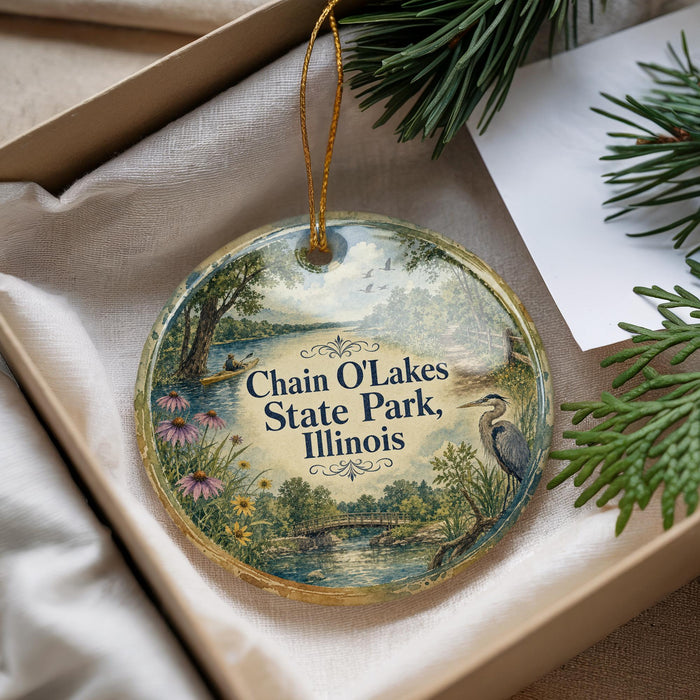 A decorative ornament with a scenic landscape of Chain O Lakes State Park, Illinois, featuring a bird and flowers, is displayed in a box with pine branches.