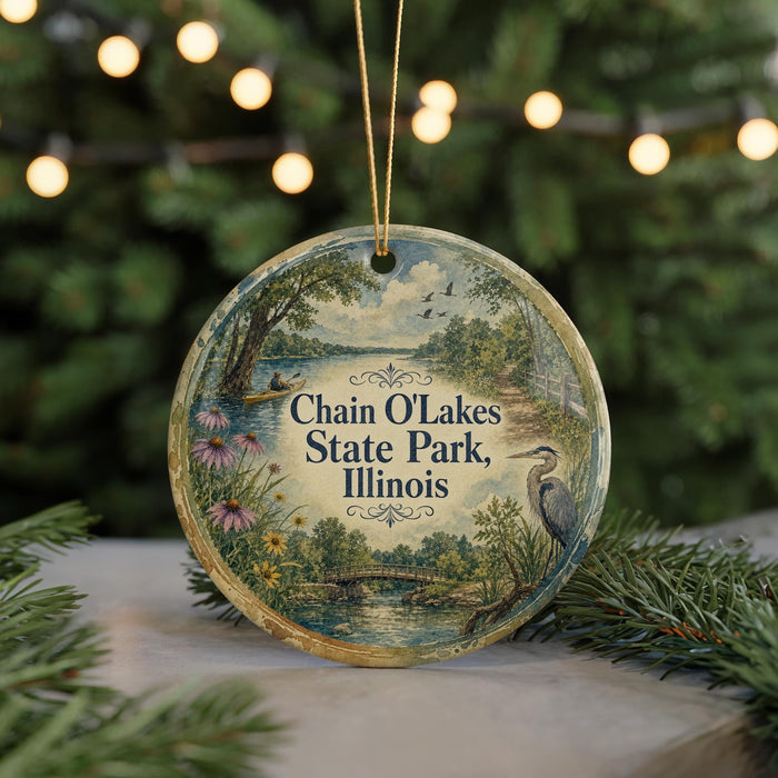 A round ornament with a scenic landscape of Chain O Lakes State Park, Illinois, featuring a bird and flowers, is displayed on a table with a blurred background of a Christmas tree and lights.