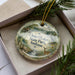 A decorative ornament in a box, featuring a scenic landscape with a deer, trees, and a body of water.