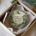 A round ornament in a box, featuring a scenic landscape with a cabin, trees, and a lake, along with a vintage-style airplane in the sky.