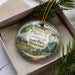 A decorative ornament in a box, featuring a scenic landscape with a cabin, trees, and a lake.