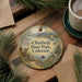A decorative ornament with a scenic landscape of Chatfield State Park, Colorado, featuring a deer, a bird, and a boat on a lake. The ornament is hanging on a wooden surface, surrounded by pine branches and a cup of coffee.