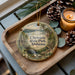A decorative ornament with a scenic image of Chewala State Park, Alabama, is displayed on a wooden tray alongside pine cones and a lit candle.