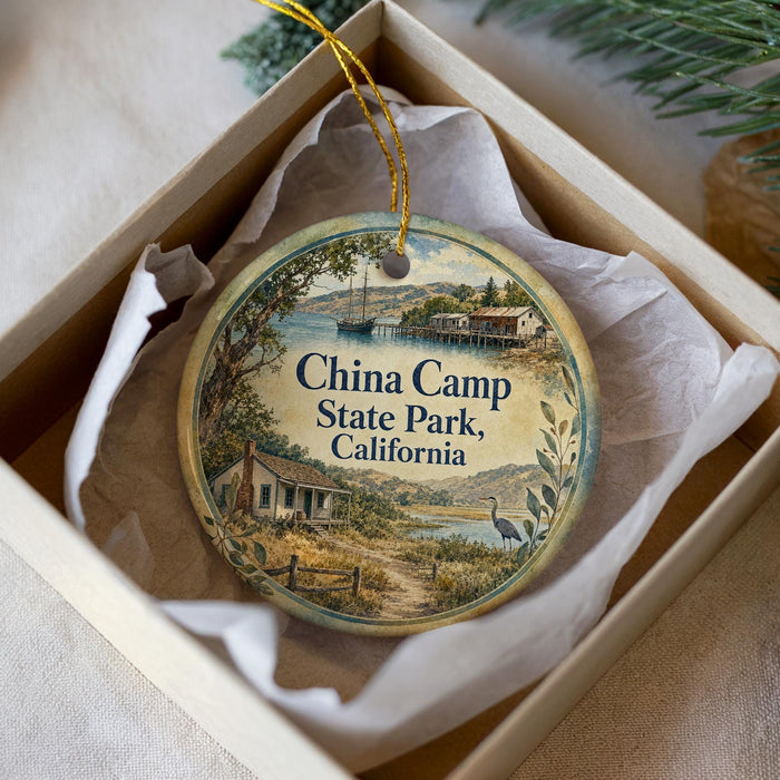 A round ornament with a scenic illustration of a coastal landscape, featuring a small house, a boat, and a bird, is displayed in a box.