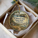 A round ornament with a scenic illustration of a coastal landscape, featuring a small house, a boat, and a bird, is displayed in a box.