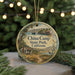 A round ornament with a scenic illustration of a coastal landscape, featuring a house, a boat, and a bird, hangs in front of a Christmas tree with twinkling lights.