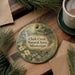 A round ornament with a scenic image of Clark Creek, surrounded by pine branches and a cup of coffee on a wooden surface.