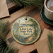A decorative ornament with a lighthouse and a lake, surrounded by pine branches and a cup of coffee on a wooden surface.