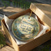 A decorative ornament in a box, featuring a lighthouse and a scenic landscape, with the text "Clear Lake State Park, Iowa" printed on it.