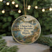 A round ornament with a scenic lake and surrounding landscape, featuring a bird and a boat, hangs in front of a blurred background with Christmas lights.