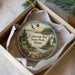 A decorative ornament in a box, featuring a scenic landscape with a lighthouse, pine trees, and a deer.