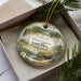 A decorative ornament in a box, featuring a scenic landscape with a castle, trees, and a body of water.