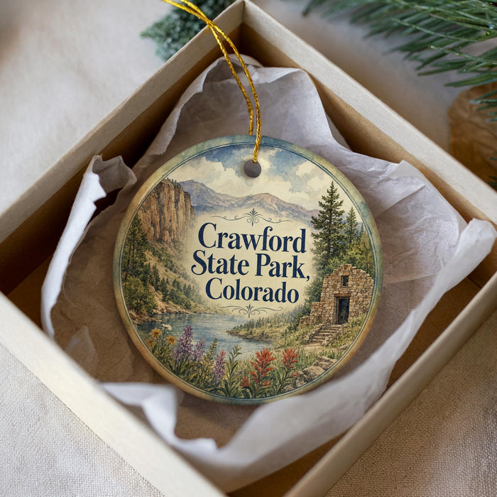 A decorative ornament in a box, featuring a scenic landscape with mountains, a river, and a small building.