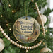A round ornament hanging on a Christmas tree, featuring a scenic landscape with mountains, a river, and a cabin.