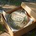 A circular ornament in a wooden box, featuring a scenic landscape with mountains, a river, and colorful flowers.