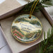 A decorative ornament with a scenic landscape of Crooked River State Park, Georgia, featuring a waterfall, pine trees, and flowers.