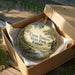 A decorative ornament in a brown box, featuring a scenic landscape with a river and trees, is placed on a wooden surface.