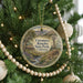 A round ornament on a Christmas tree, featuring a scenic landscape with a windmill and a river, surrounded by greenery and twinkling lights.