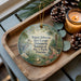 A decorative ornament featuring a bird and tropical plants, placed on a wooden tray next to a lit candle and pine cones.
